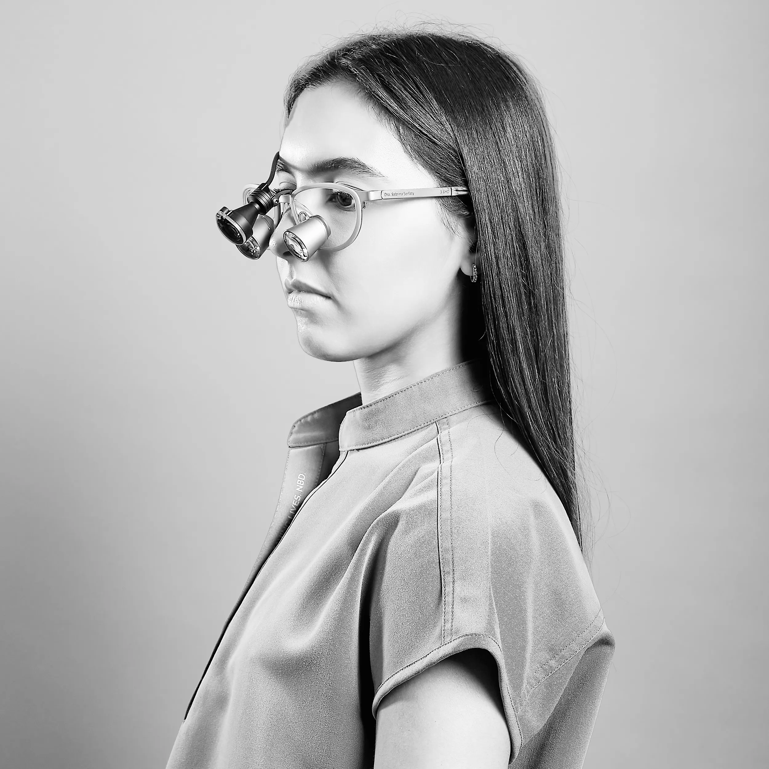 Profile studio portrait in black and white of a woman with long dark hair wearing translucent glasses, photographed against a soft background.