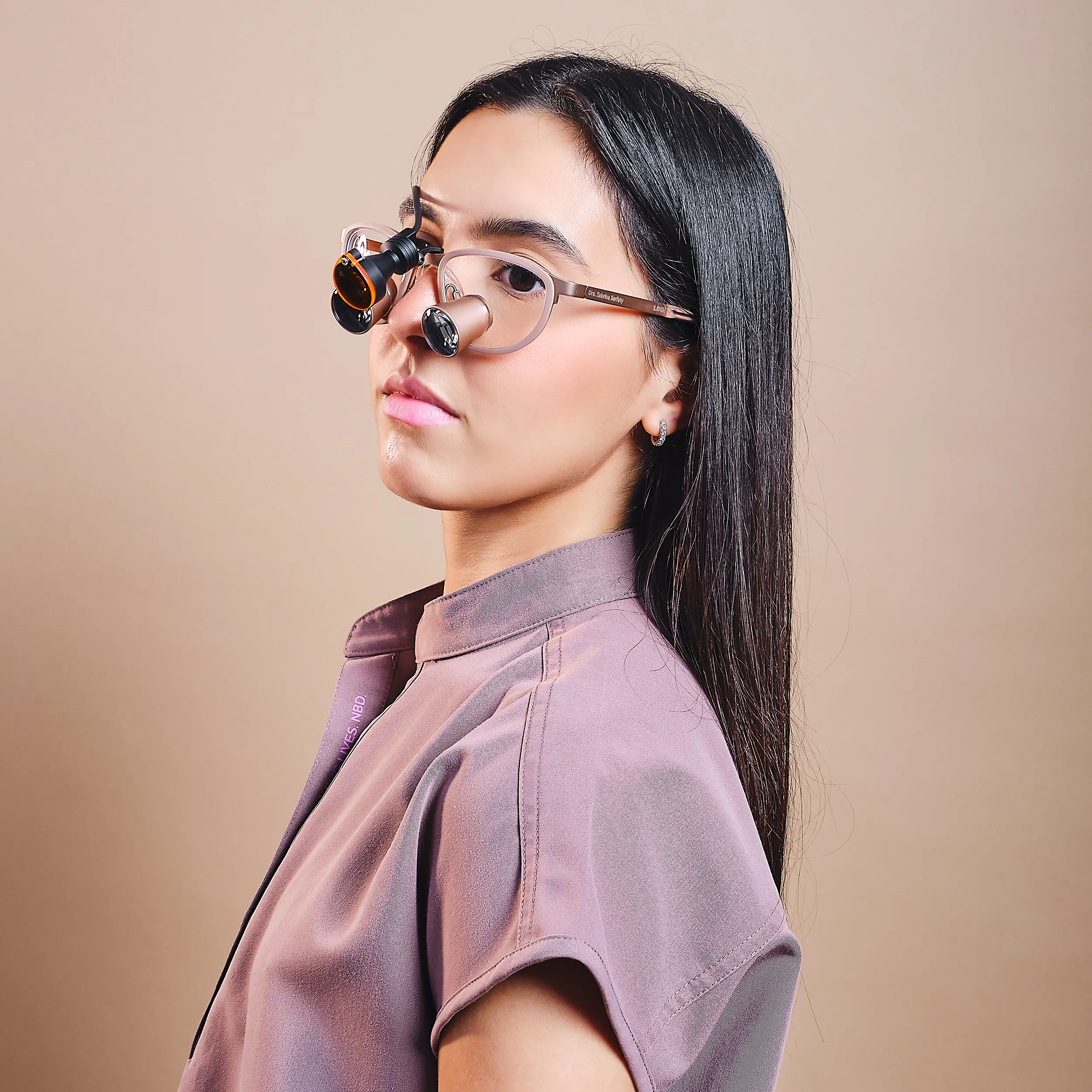 Studio portrait of a woman with long dark hair wearing translucent glasses, photographed against a soft beige background.