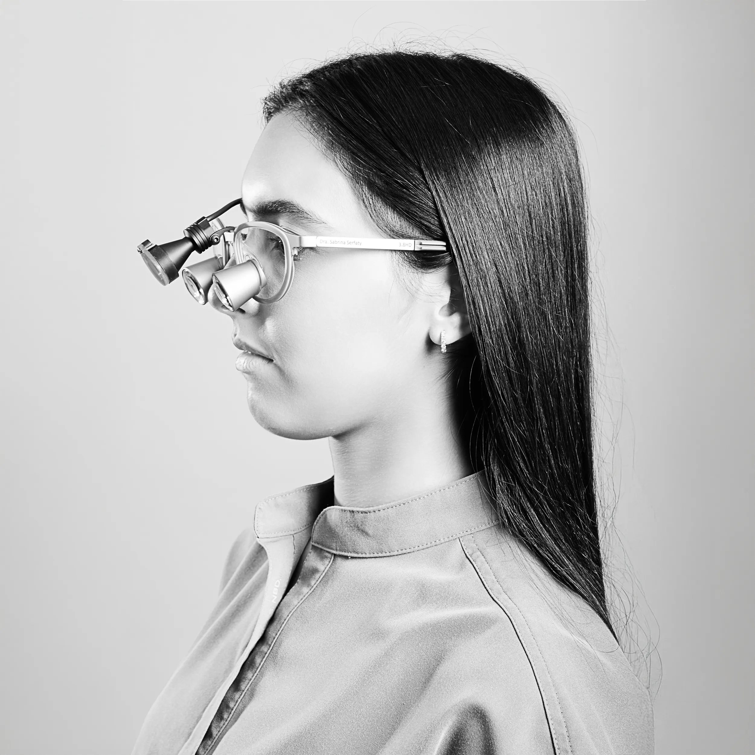 Profile studio portrait in black and white of a woman with long dark hair wearing translucent glasses, photographed against a soft background.
