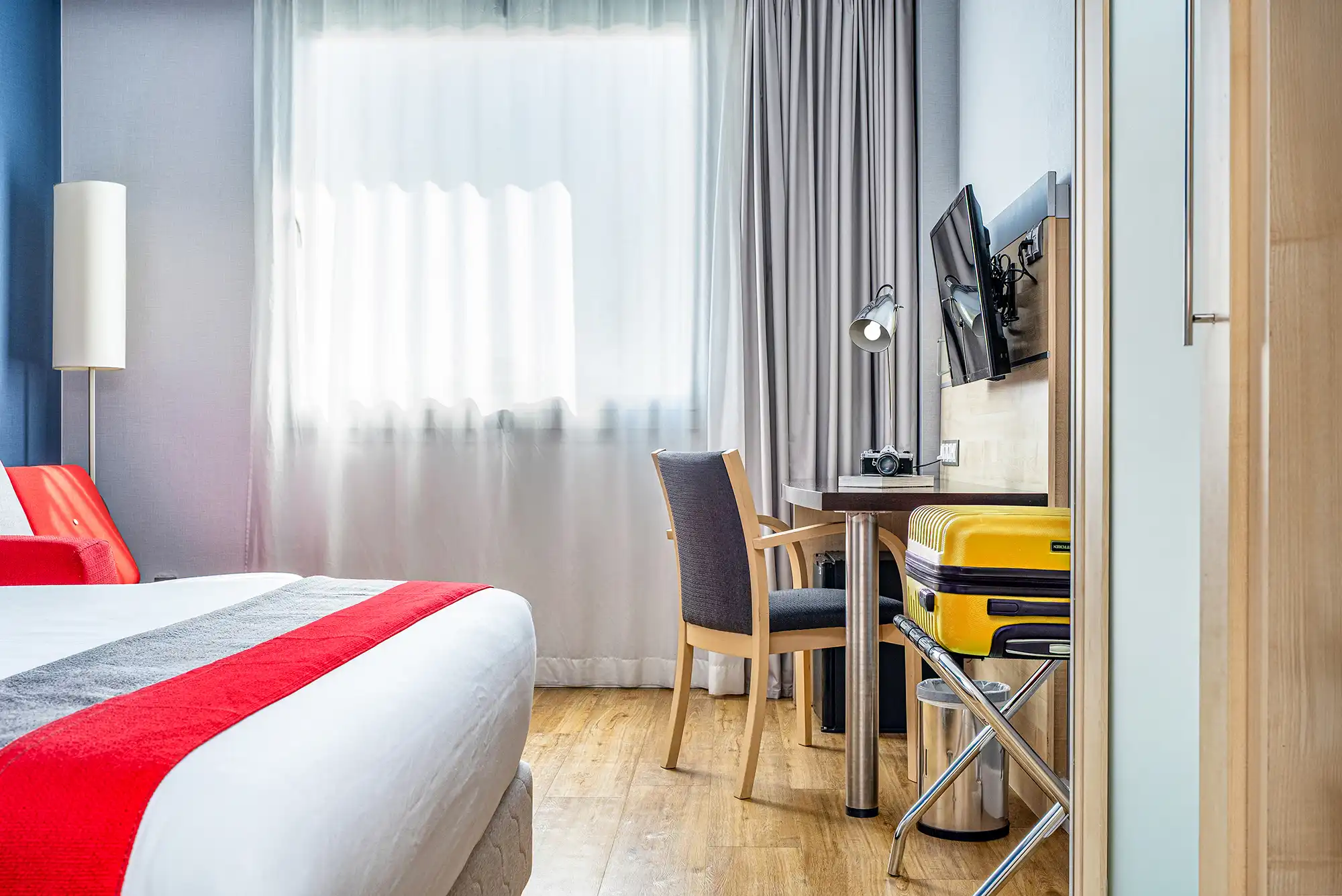 Habitación de hotel luminosa con cama, escritorio, televisión y grandes ventanales.