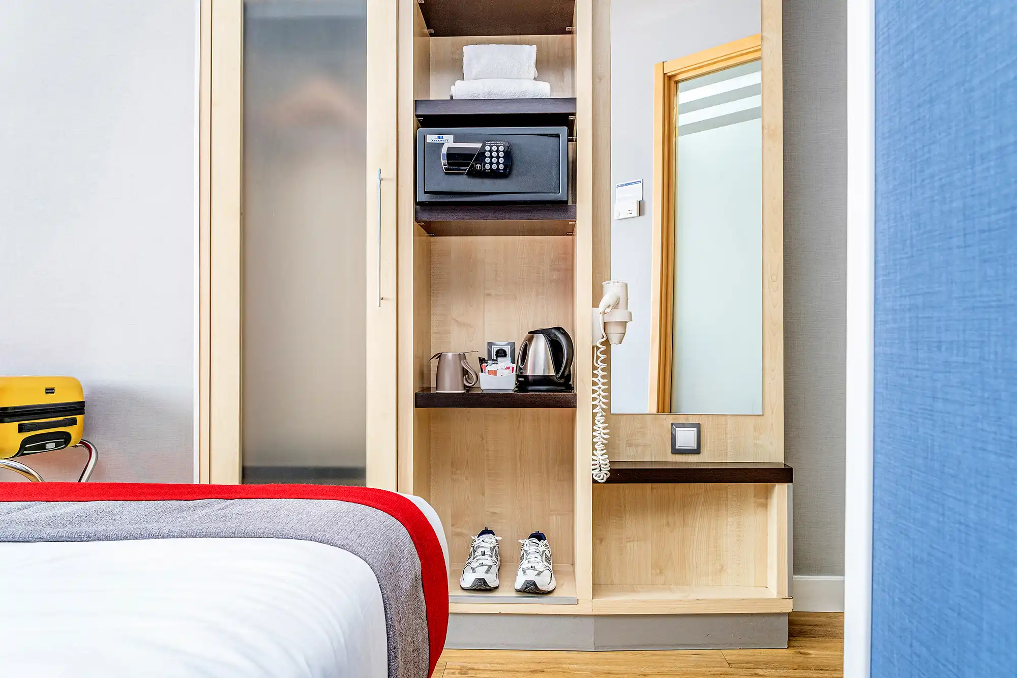 Habitación de hotel con cama, armario abierto, caja fuerte, espejo y espacio para guardar el equipaje.