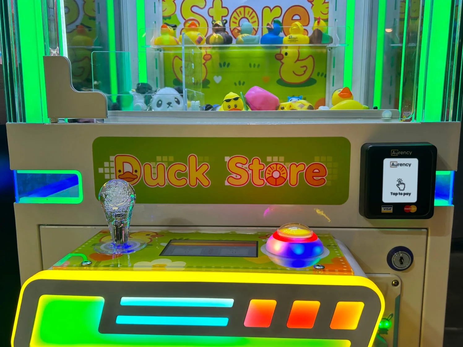 A neon Duck Store claw machine with an Aurency A250 reader mounted on the cabinet showing Tap to pay