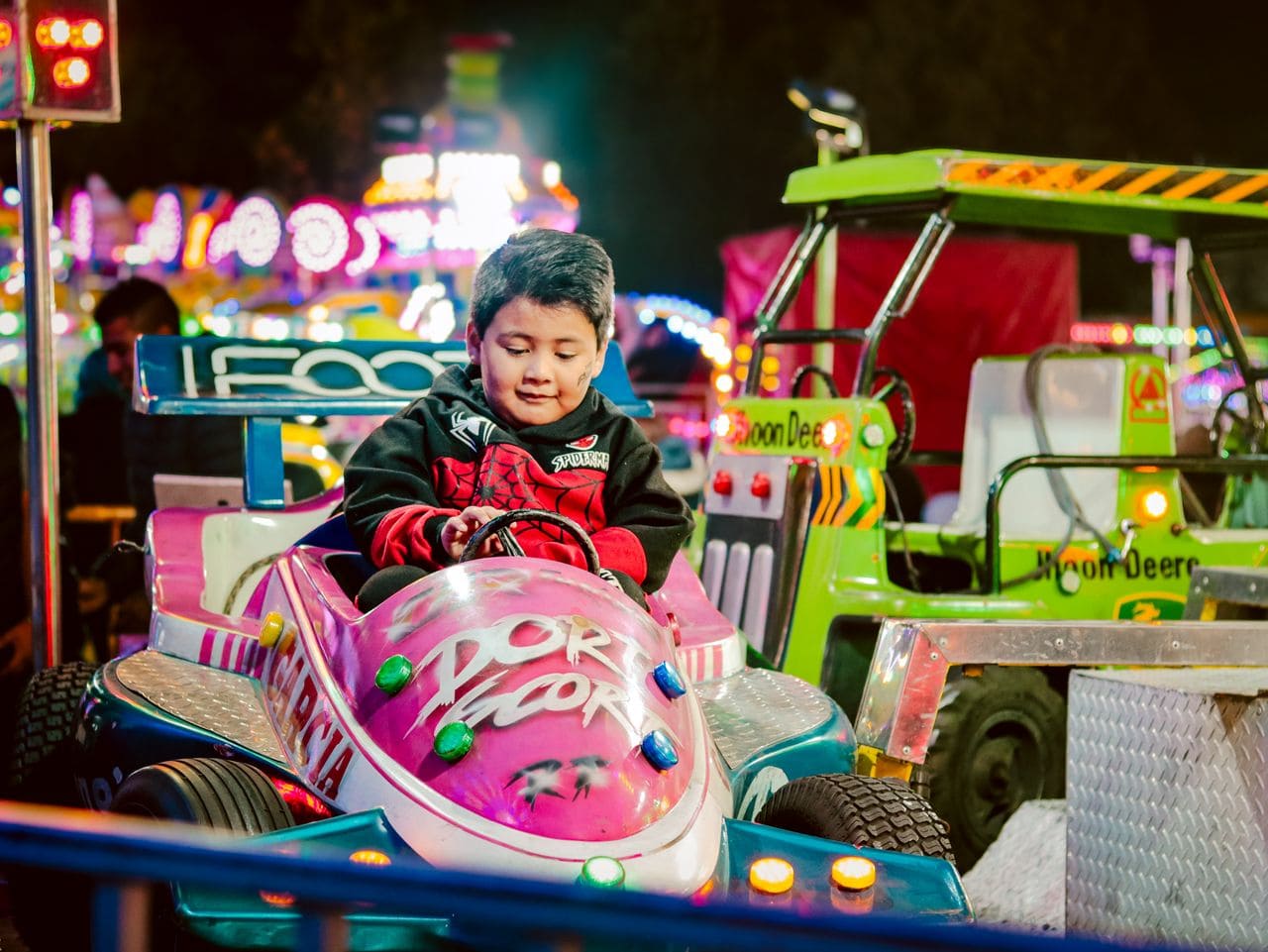 Un bambino piccolo su un go-kart rosa in un parco divertimenti notturno, con coloratissime giostre da sala giochi sullo sfondo