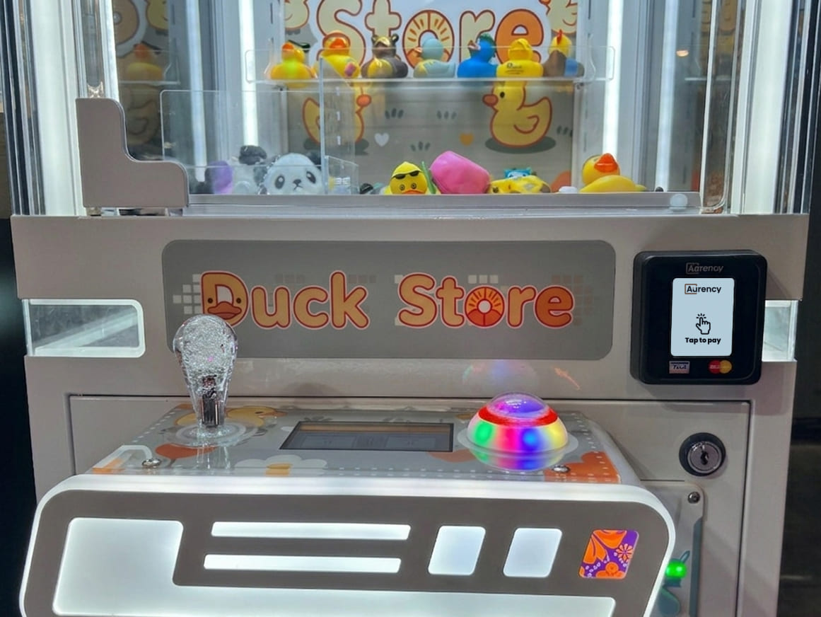 An Aurency A250 contactless reader mounted on a Duck Store claw machine filled with rubber ducks and plush toys