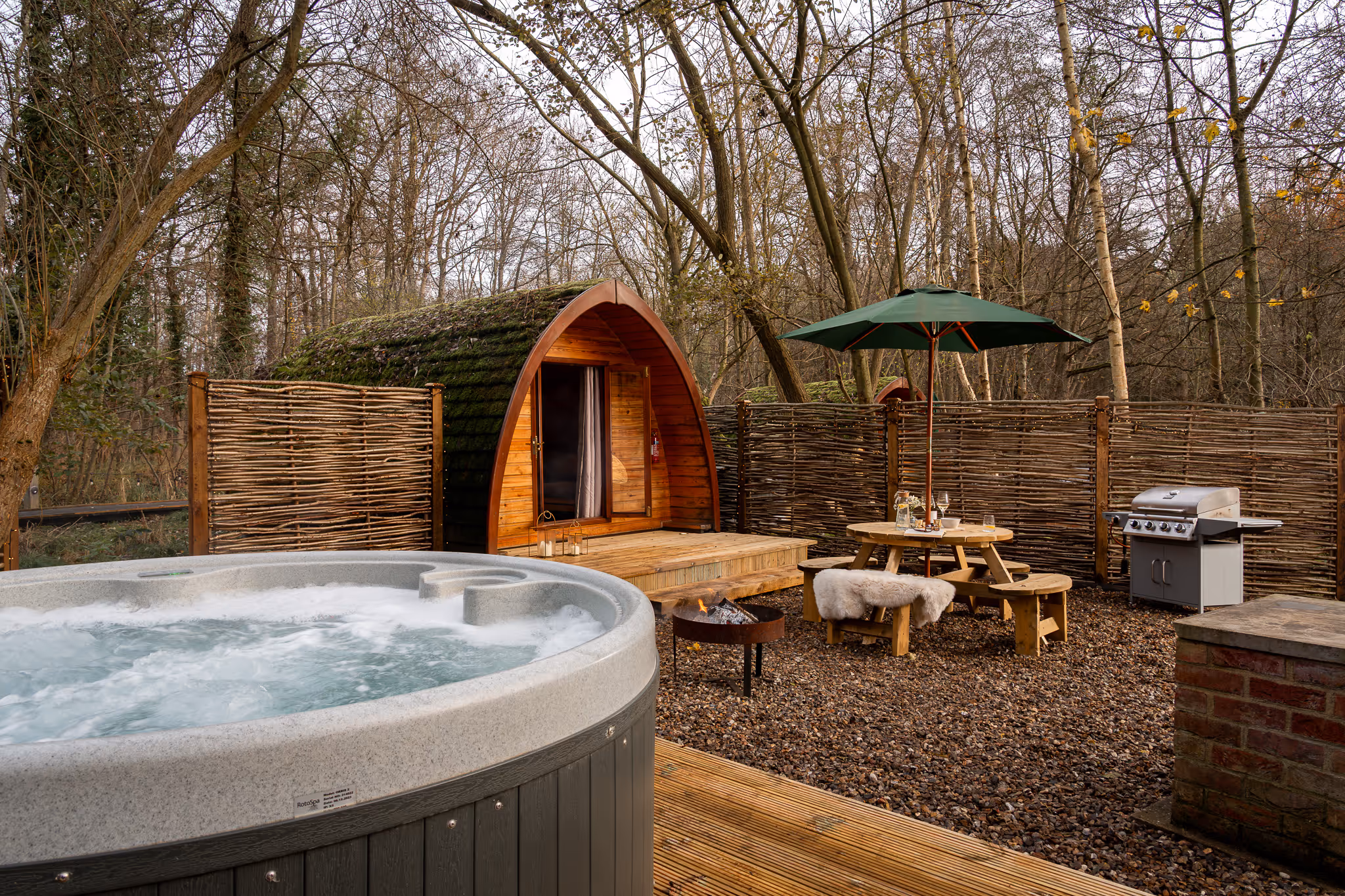 Outdoor glamping setup with a wooden pod, bubbling hot tub, fire pit, picnic table with green umbrella, and a barbecue grill in a wooded area.