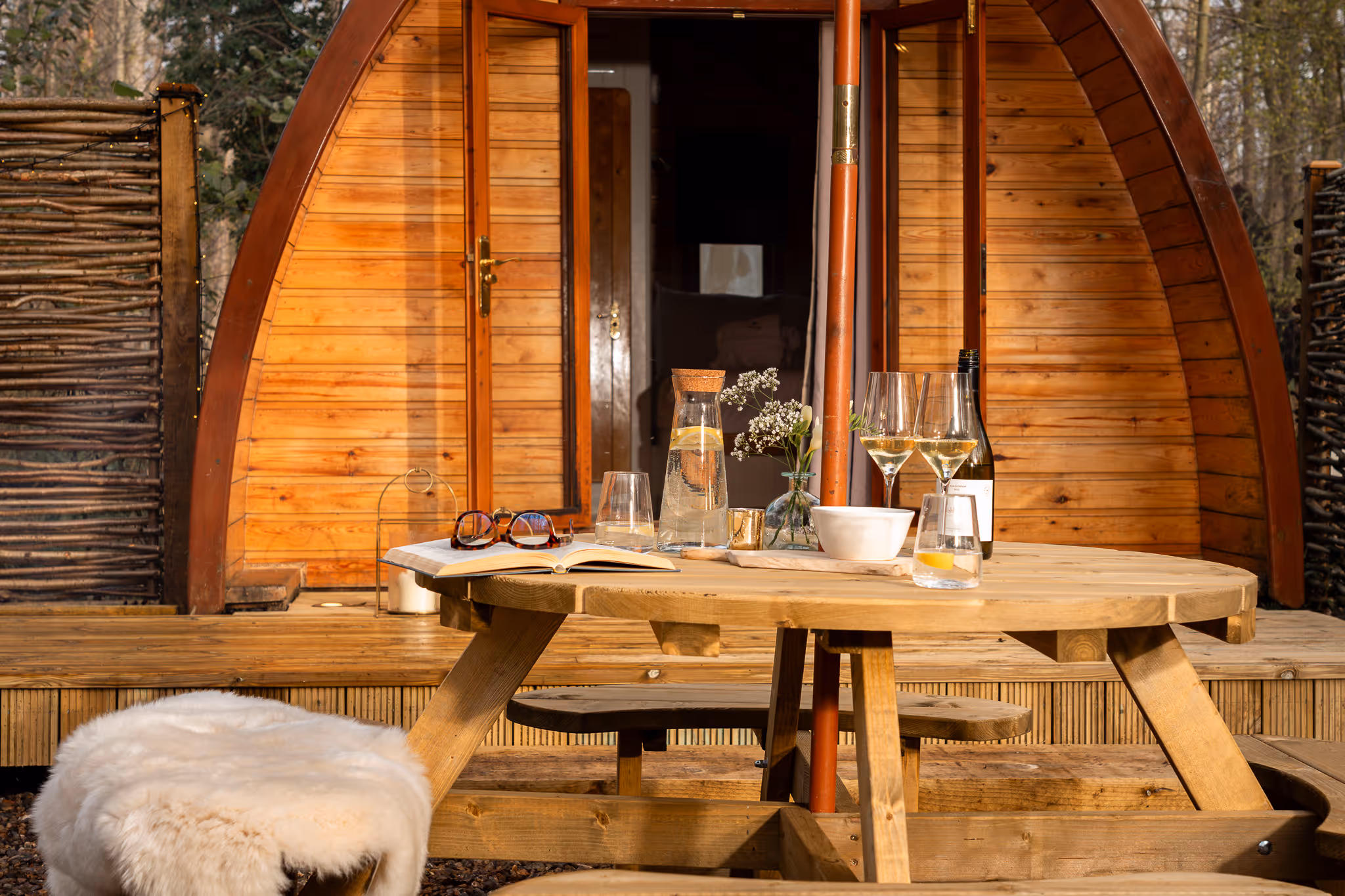 Round wooden picnic table with glasses, a carafe of water with lemon, a wine bottle, and an open book in front of a wooden pod cabin with open doors.
