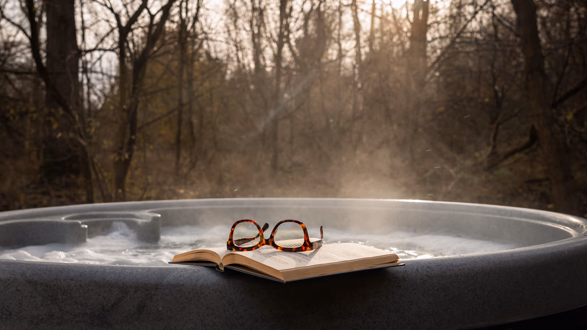 Open book with tortoiseshell eyeglasses resting on the edge of a steaming hot tub in a wooded area.