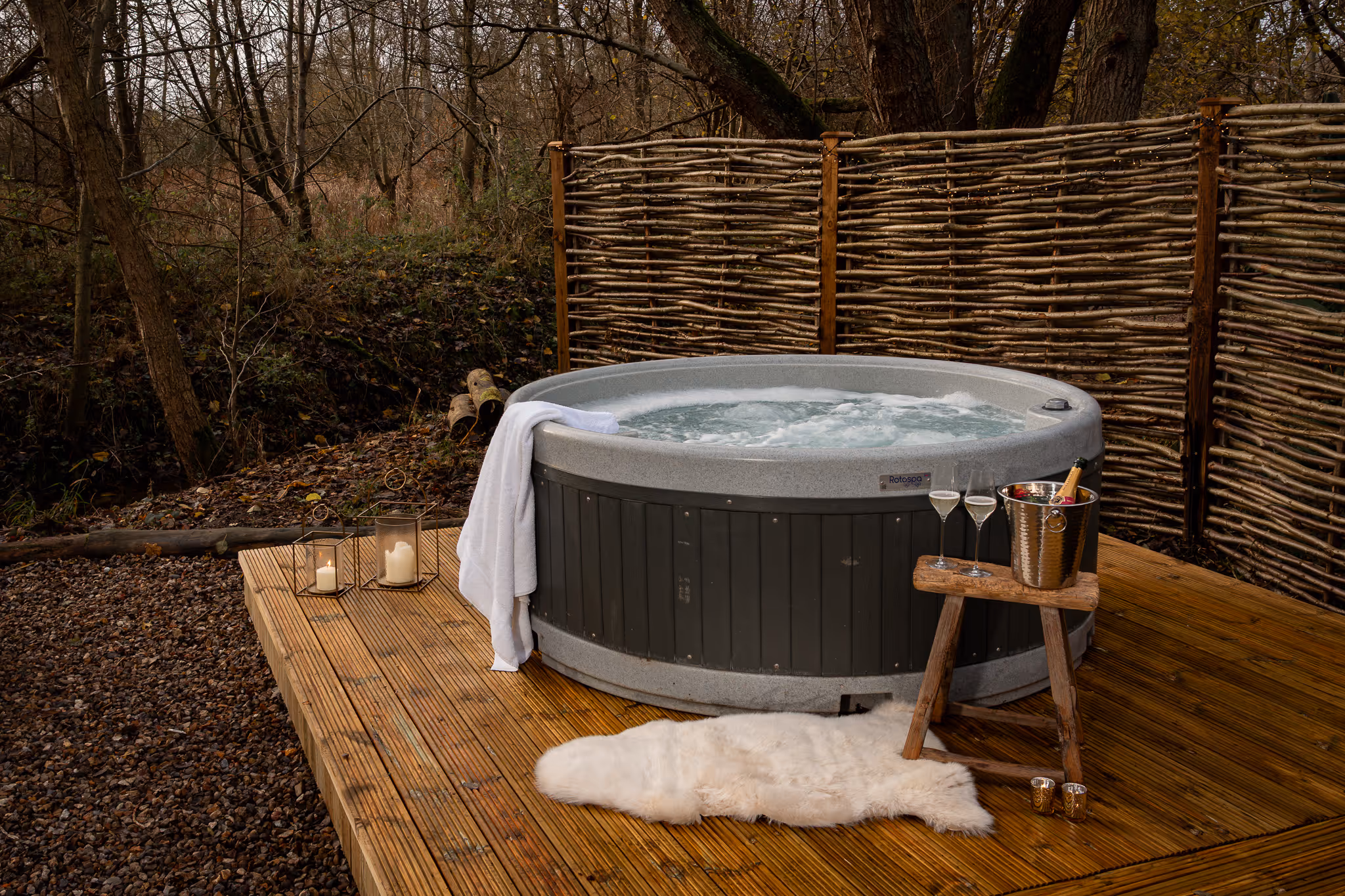 Outdoor hot tub on wooden deck with white towel, candles, champagne bucket, and glasses, surrounded by wicker privacy fencing and forest.