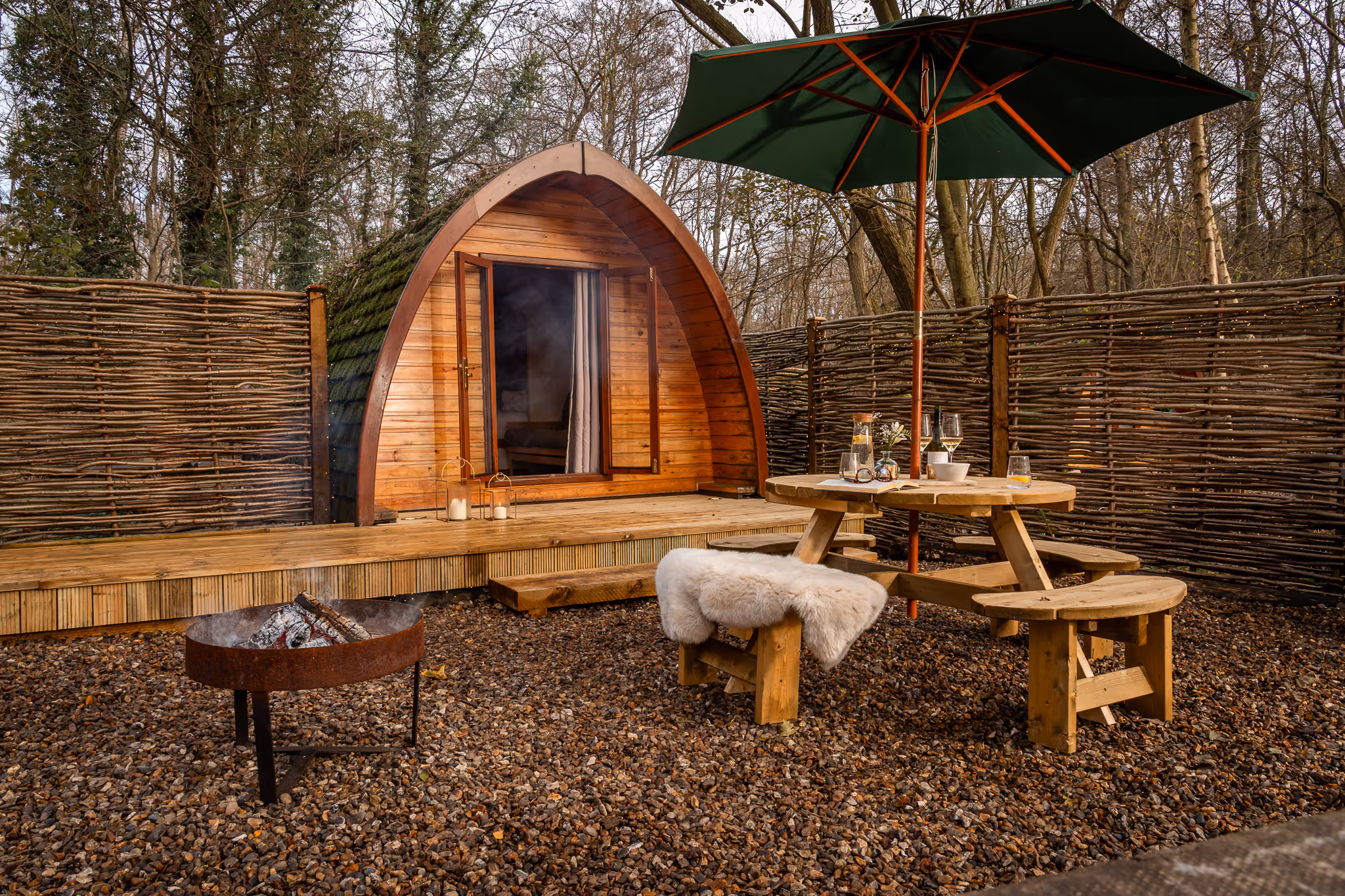 Wooden pod cabin with open doors, a fire pit, and a round picnic table with benches and a green umbrella in a fenced outdoor area covered with gravel.