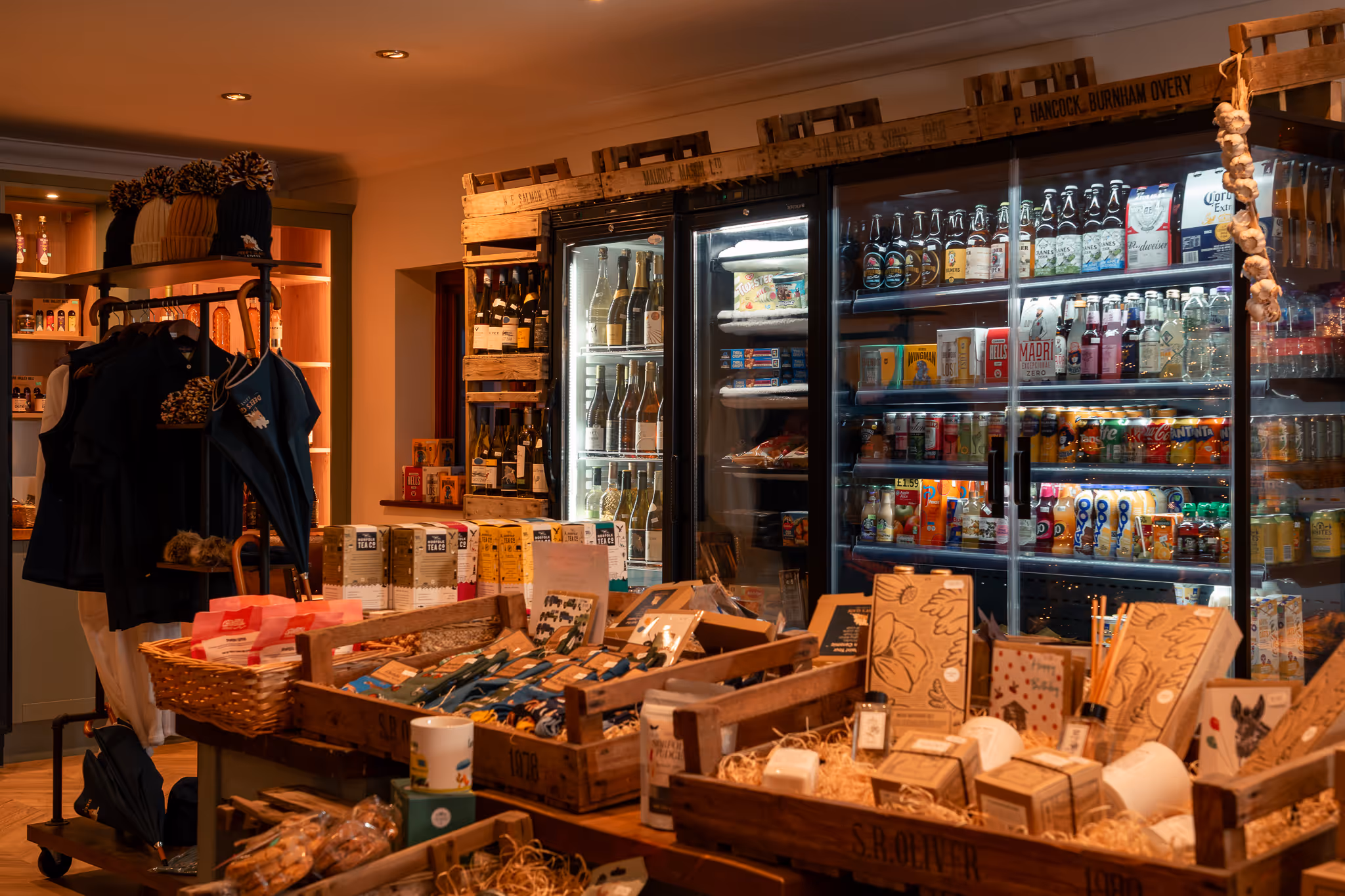 Cozy shop interior with wooden crates displaying assorted gift items, clothing, and refrigerated shelves stocked with beverages and snacks.