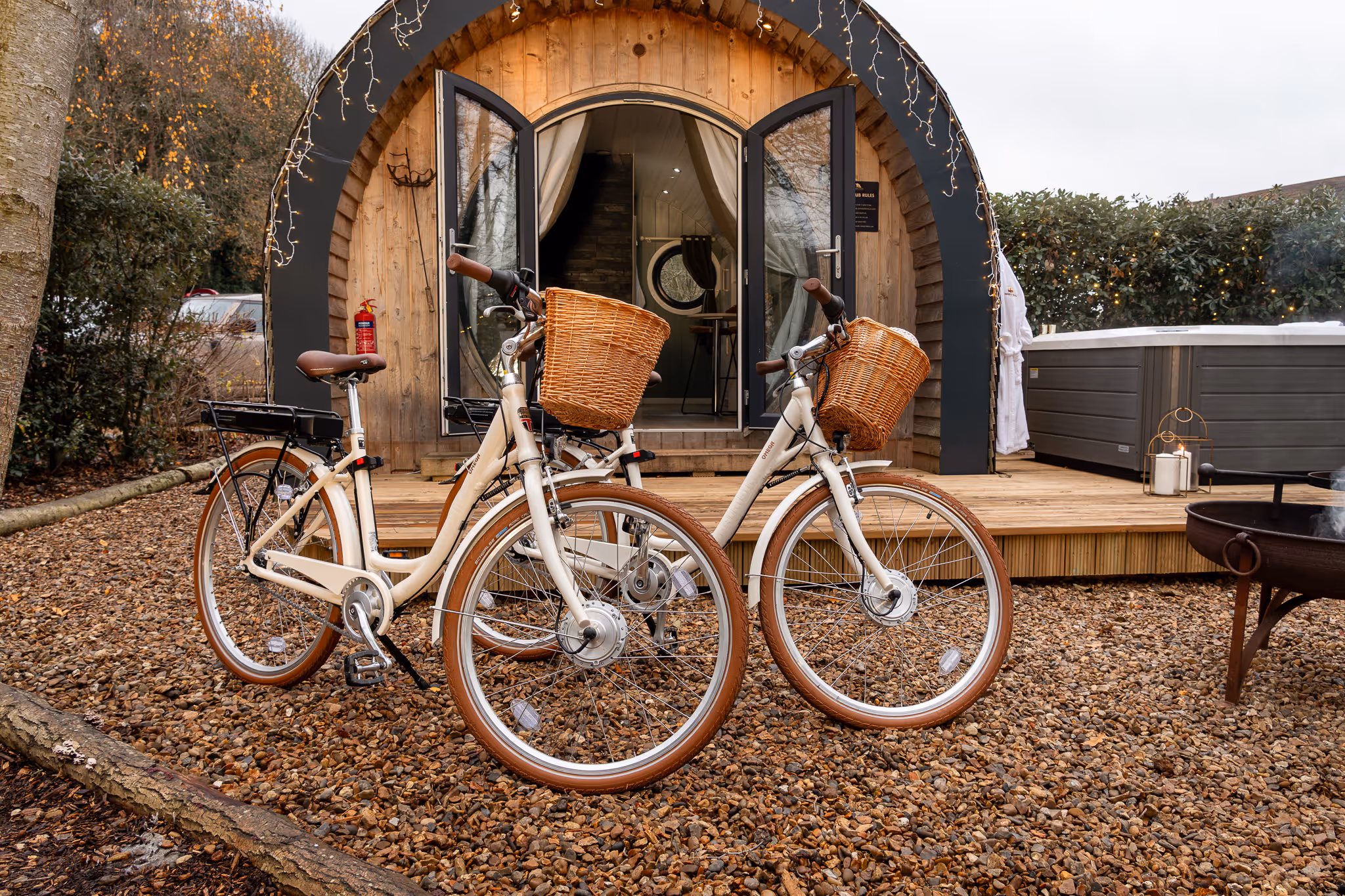 Two cream-colored electric bicycles with wicker baskets parked on gravel outside a wooden glamping pod with open doors and string lights.