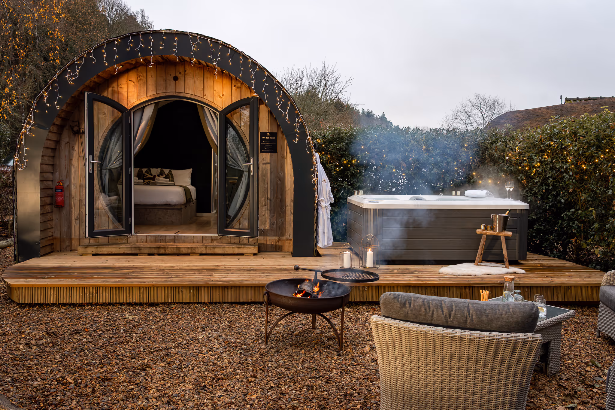 Cozy outdoor retreat with a curved wooden cabin featuring open double doors revealing a bed, a steaming hot tub on a wooden deck, a fire pit, and wicker seating.