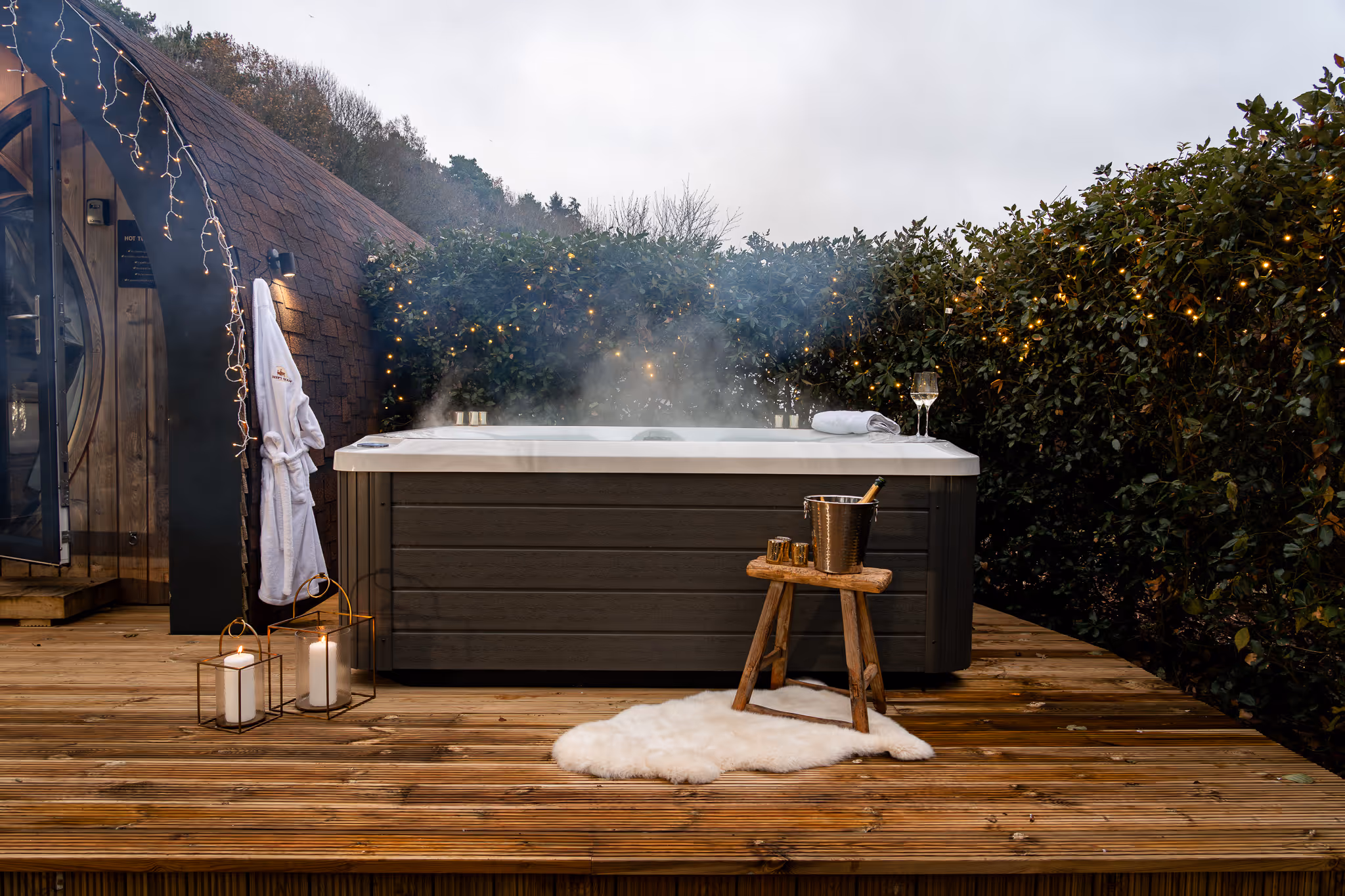 Steaming outdoor hot tub on wooden deck surrounded by greenery, with a wooden stool holding a champagne bucket and glasses nearby.