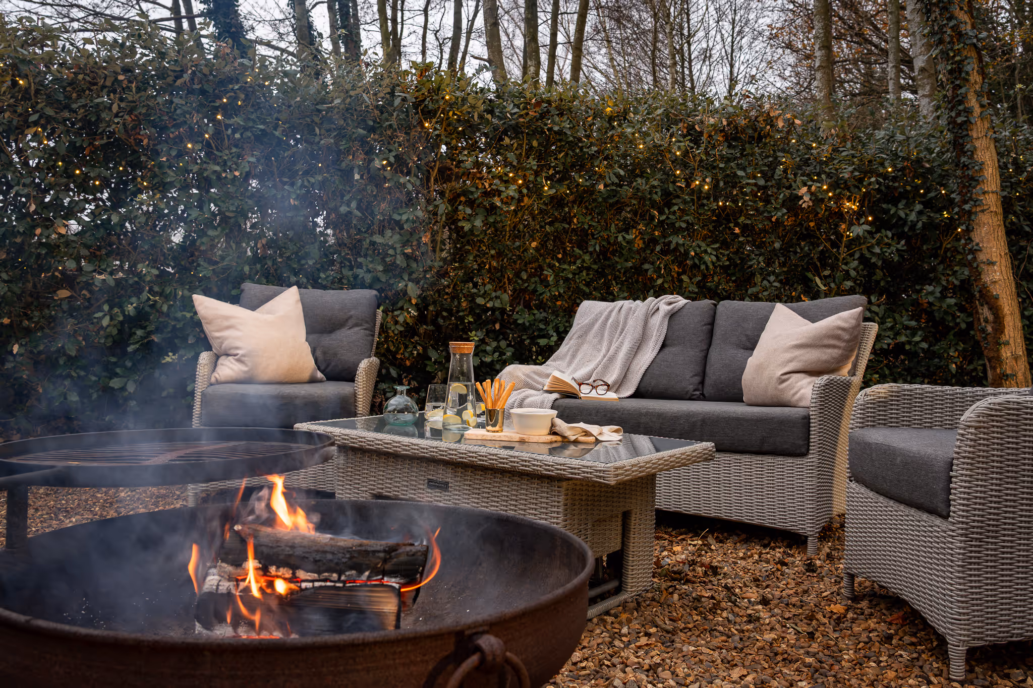 Outdoor seating area with gray wicker sofa and chairs, beige pillows, a coffee table with drinks and snacks, a blanket, and a fire pit with burning logs.