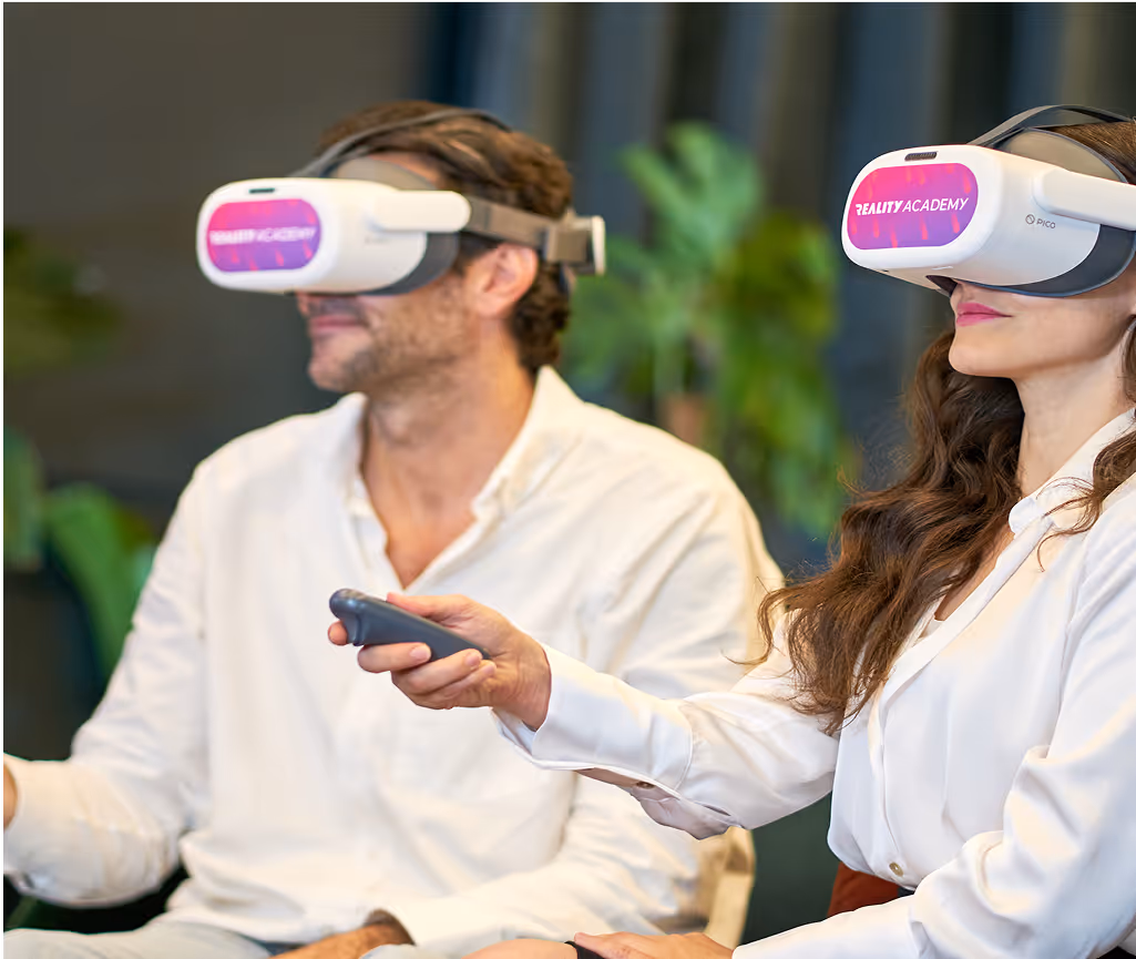 Un homme et une femme portent des casques de réalité virtuelle marqués “REALITY ACADEMY” et tiennent des manettes, plongés dans une expérience immersive. Ils sont assis côte à côte dans un environnement intérieur moderne avec des plantes en arrière-plan.