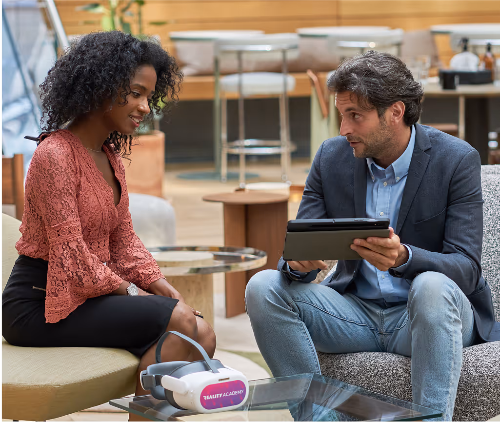 Un homme et une femme discutent assis dans un espace moderne, avec une tablette en main. Sur la table devant eux se trouve un casque de réalité virtuelle portant l’inscription “REALITY ACADEMY”.