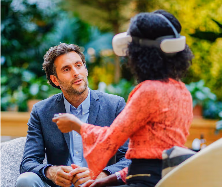 Une femme portant un casque de réalité virtuelle interagit avec un homme en costume qui l’observe attentivement. Ils sont assis dans un espace intérieur moderne et verdoyant, évoquant un cadre de formation professionnelle ou d’entretien immersif.