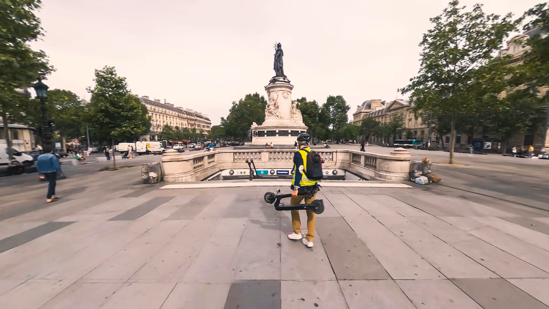 Un utilisateur de trottinette plie son engin pour traverser une zone piétonne près d’une station de métro, illustrant le module « Les comportements pour rouler sereinement à trottinette » qui enseigne les bonnes pratiques pour partager l’espace urbain en toute sécurité.