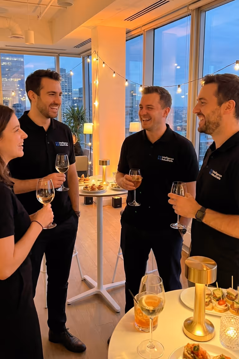 Team evening in the office overlooking the city: colleagues talking and toasting around high tables, warm and friendly atmosphere.