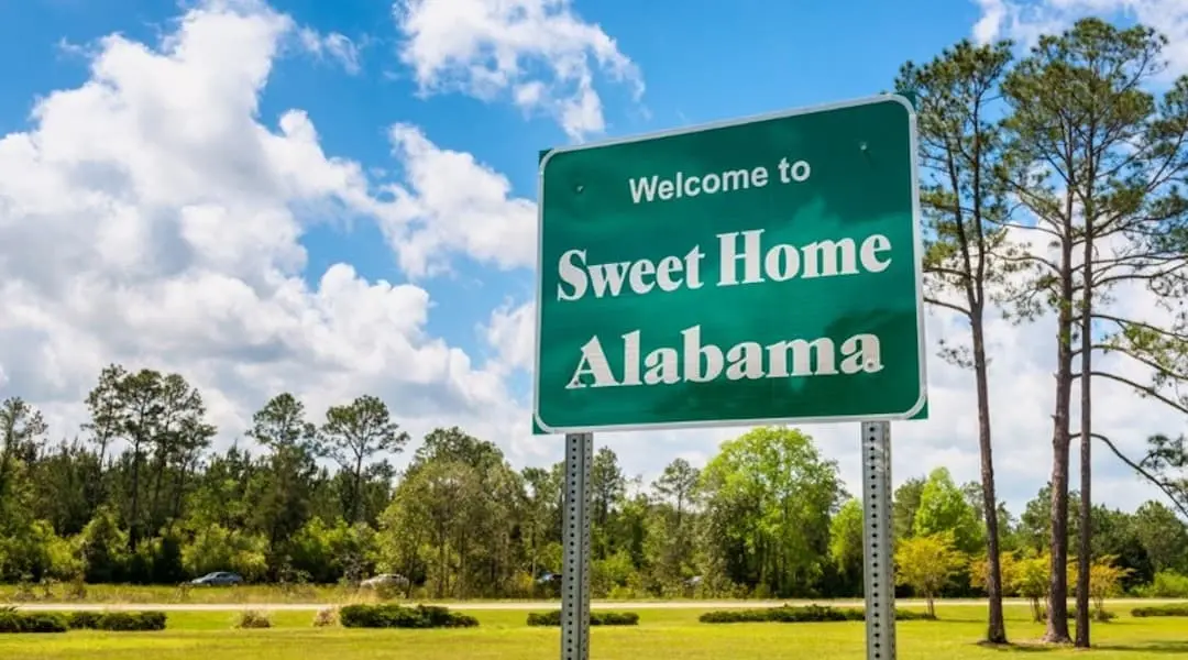 Welcome to Sweet Home Alabama roadside sign along a highway with trees, blue sky, and clouds in the background.