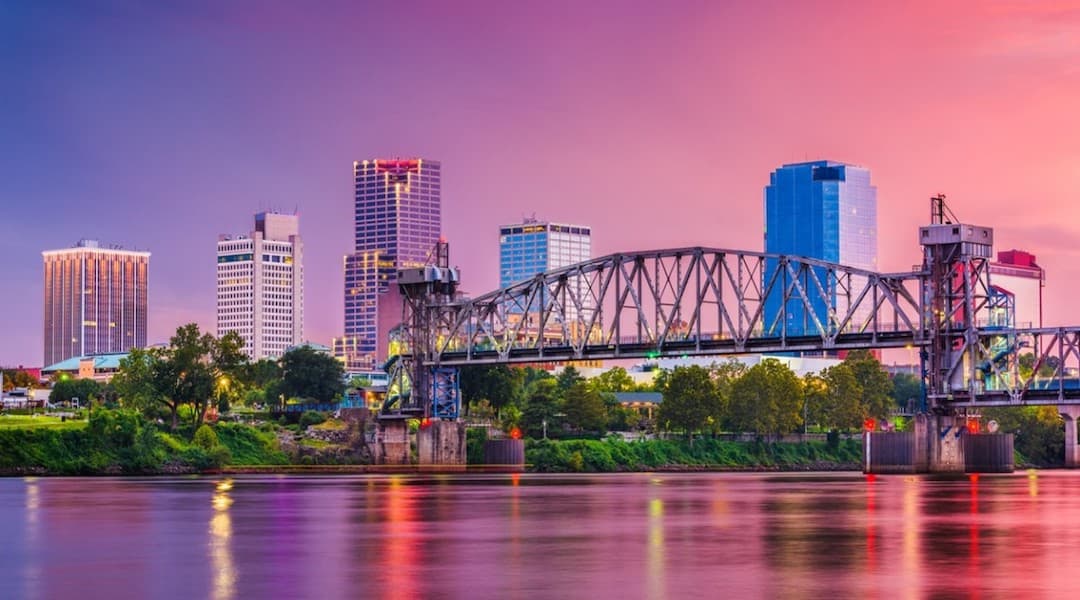Little Rock Arkansas skyline at dusk, featuring the Arkansas River and a truss bridge with illuminated downtown buildings.