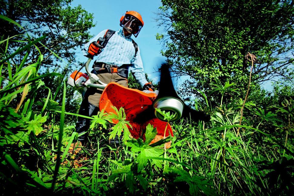 Man wearing protective gear using a grass cutter to trim tall green brush in a sunny outdoor setting.