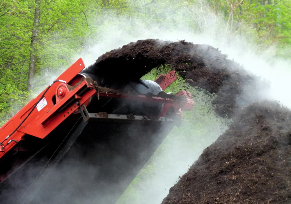 A compost grinder machine is pouring a steaming pile of rich, dark compost in a green outdoor setting.