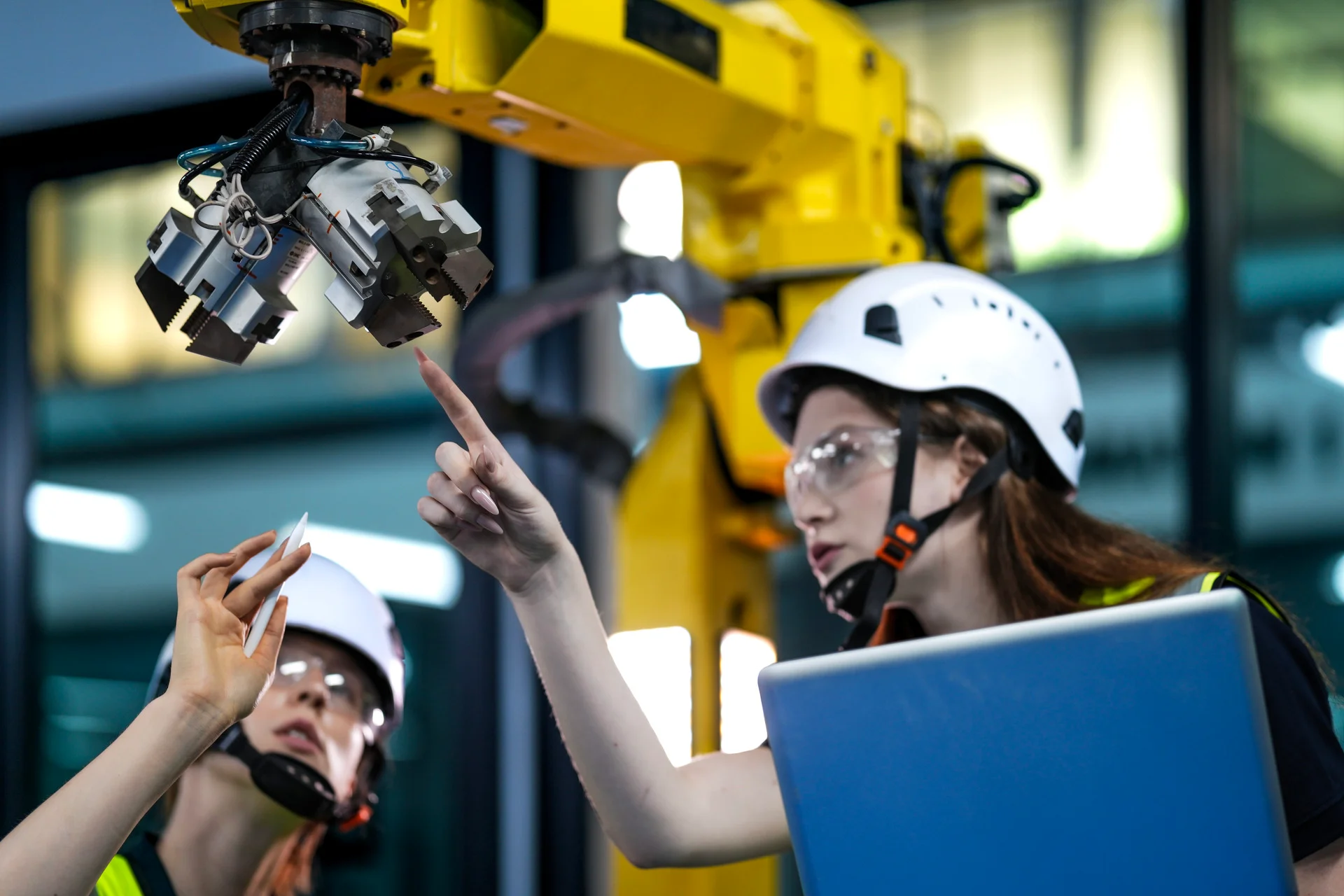 Two frontline engineers, wearing hard hats and safety glasses, collaborate in a factory setting. One engineer uses a stylus and points to a component on a large yellow robotic arm, while the other holds a laptop and points to a corresponding detail on the screen, illustrating real-time execution verification.