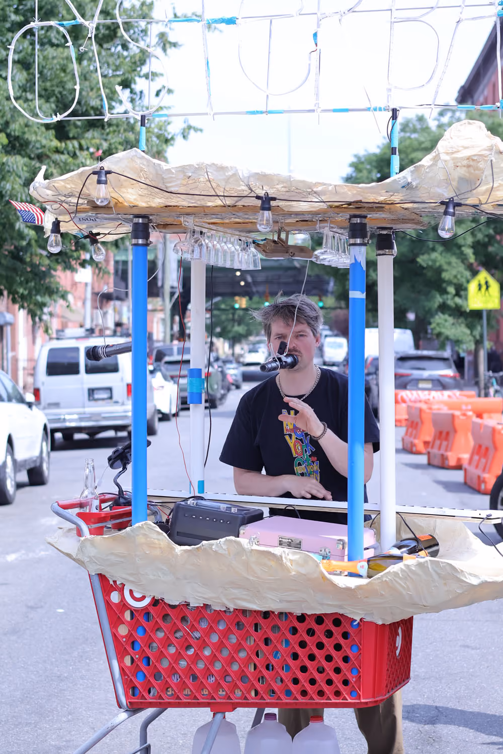 Oyster Piano Bar: a person sings into a microphone behind a red shopping cart topped with a canopy and shell-like rim, on a city street.
