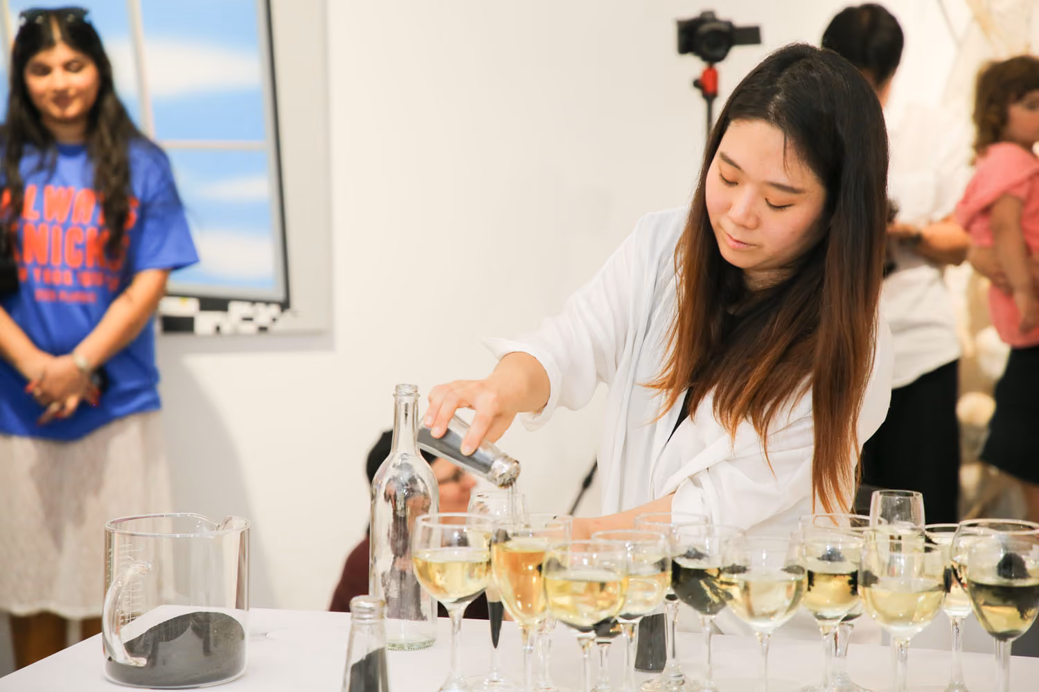 Our Oily and Lush Vitality: a woman in white pours into a row of wine glasses while a visitor in a blue T-shirt watches beside a framed sky painting.
