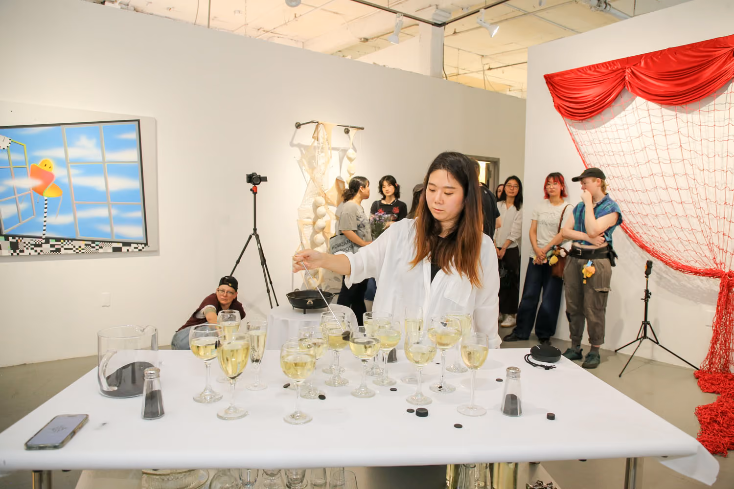 Installation view of Our Oily and Lush Vitality: a woman in white tends a long table of wine glasses as onlookers stand near a red netted curtain and a painting.