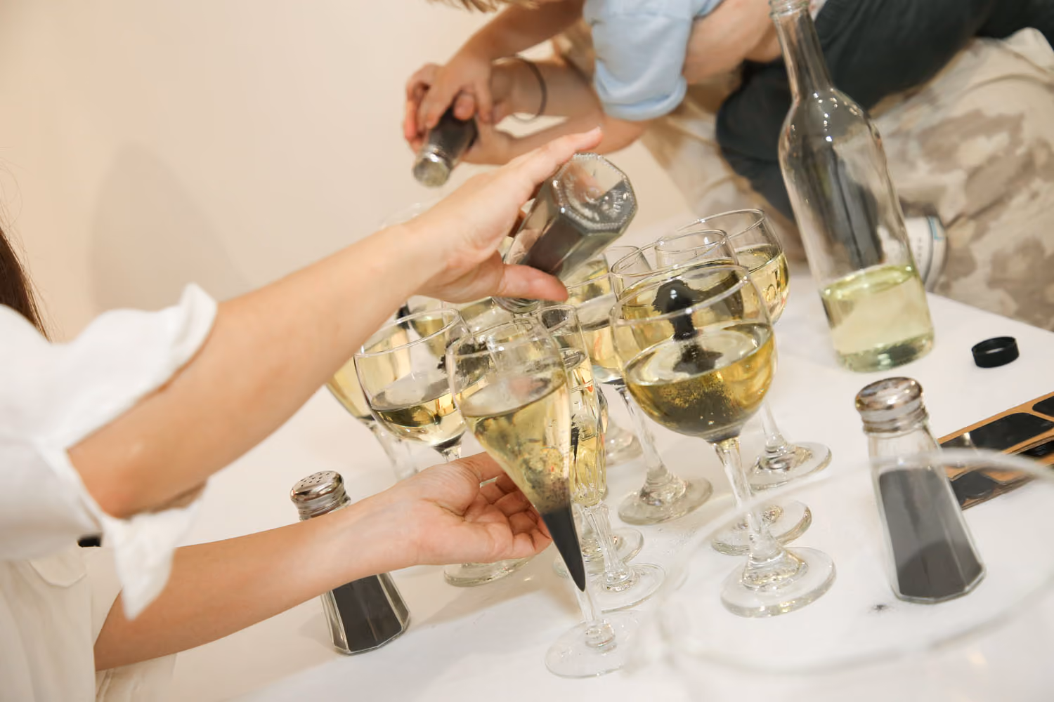 Our Oily and Lush Vitality: hands pour dark powder from small shakers into wine glasses of pale wine on a white table.