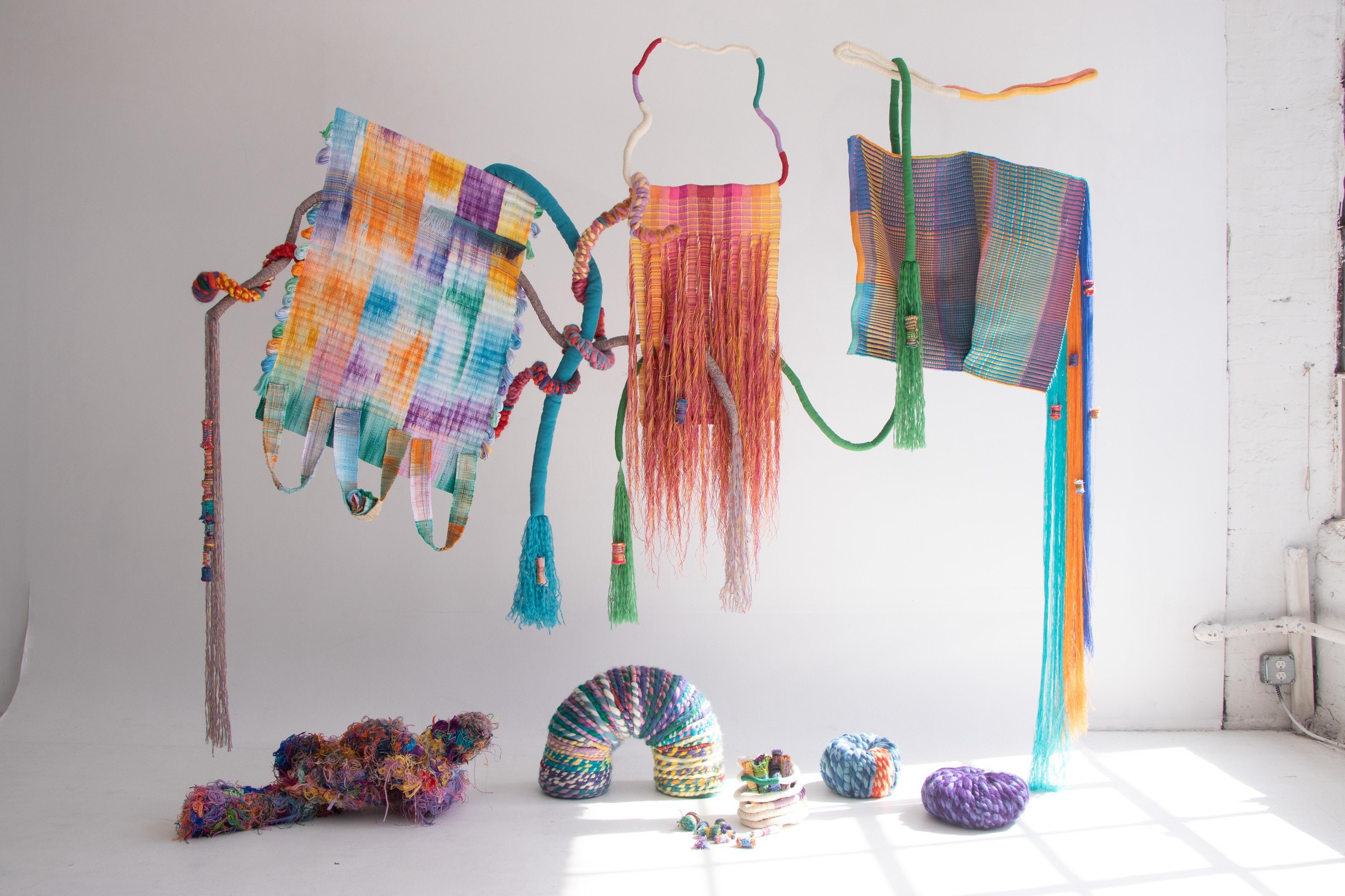 Lauren Colbert, weak ties, 2023: hanging woven textiles in rainbow, orange, and teal, with tassels and small knotted forms on the floor of a white room.