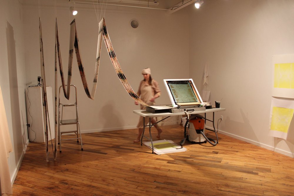 Work by Sirkku Ketola: installation of long printed fabric strips hung in a zigzag pattern, there is a person walking behind a screen printing table.
