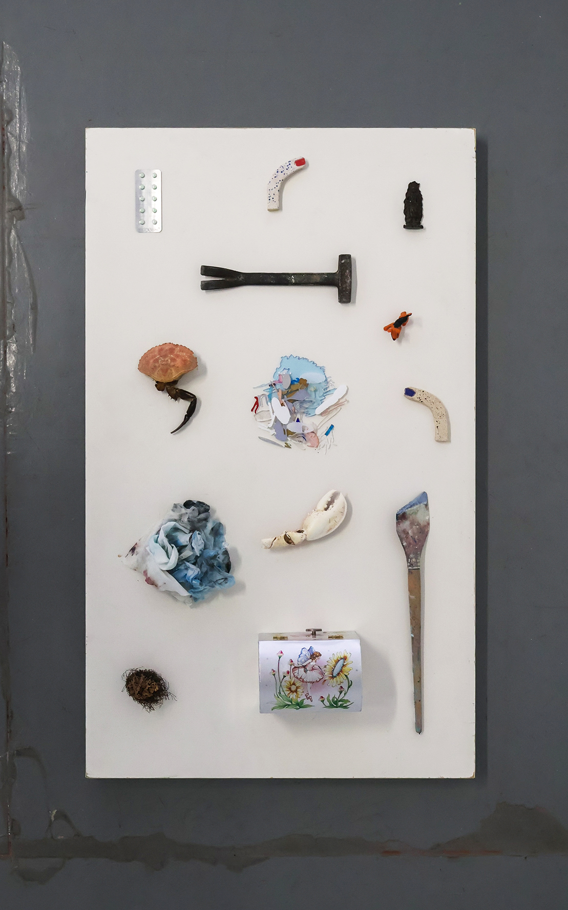 Collected Objects from Season III, 2026 Artists-in-Residence: small found objects, including a hammer, shells, a brush, and a painted tin, arranged on a white board against a dark wall.