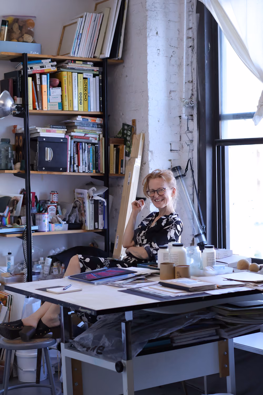 NARS Spring Open Studios 2026: a smiling woman sits at a drafting table in a studio with tall bookshelves and a window behind her.