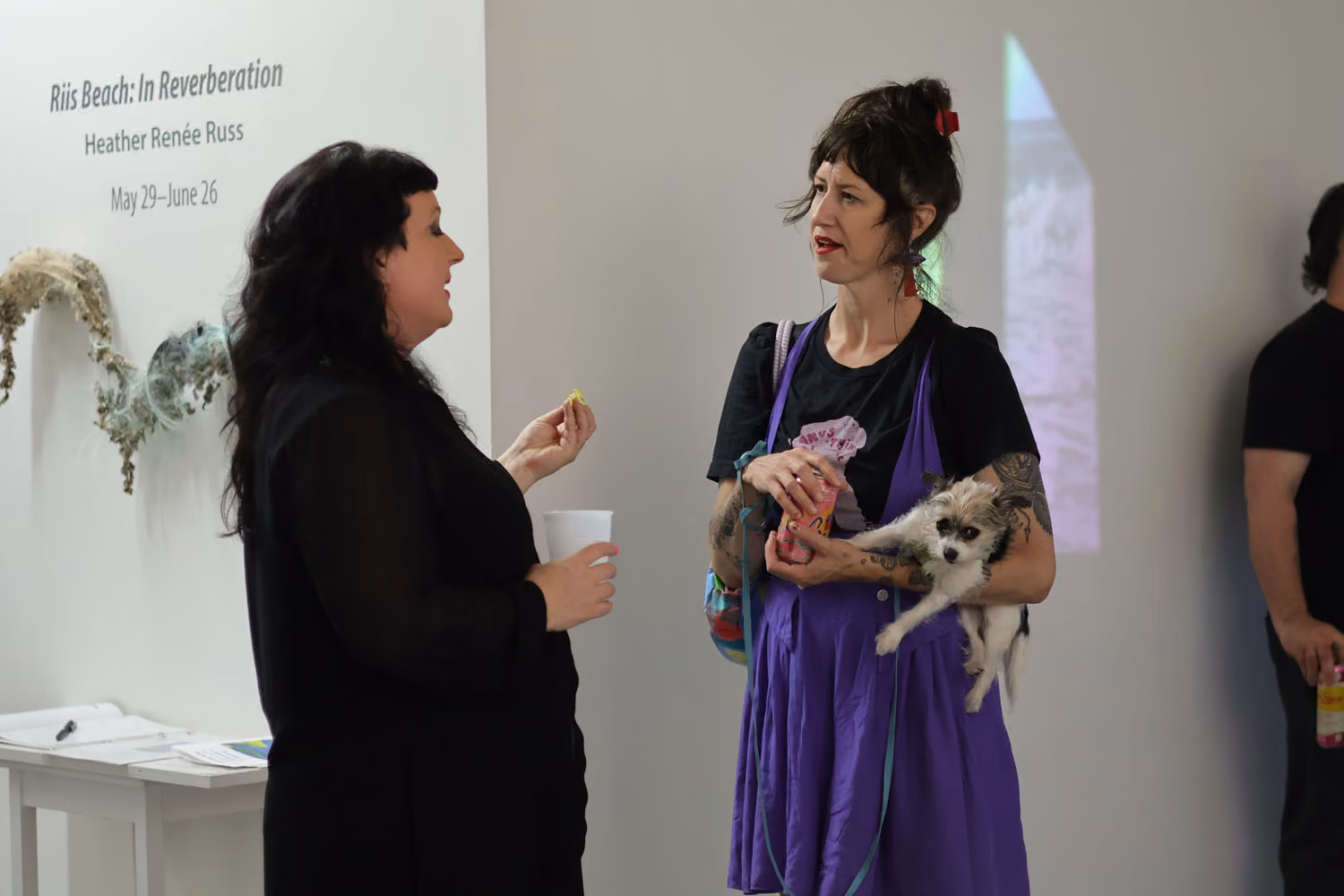NARS Spring Open Studios 2026: two women talk, one holding a small dog, beside a wall with exhibition title text and a sculpture on the left.