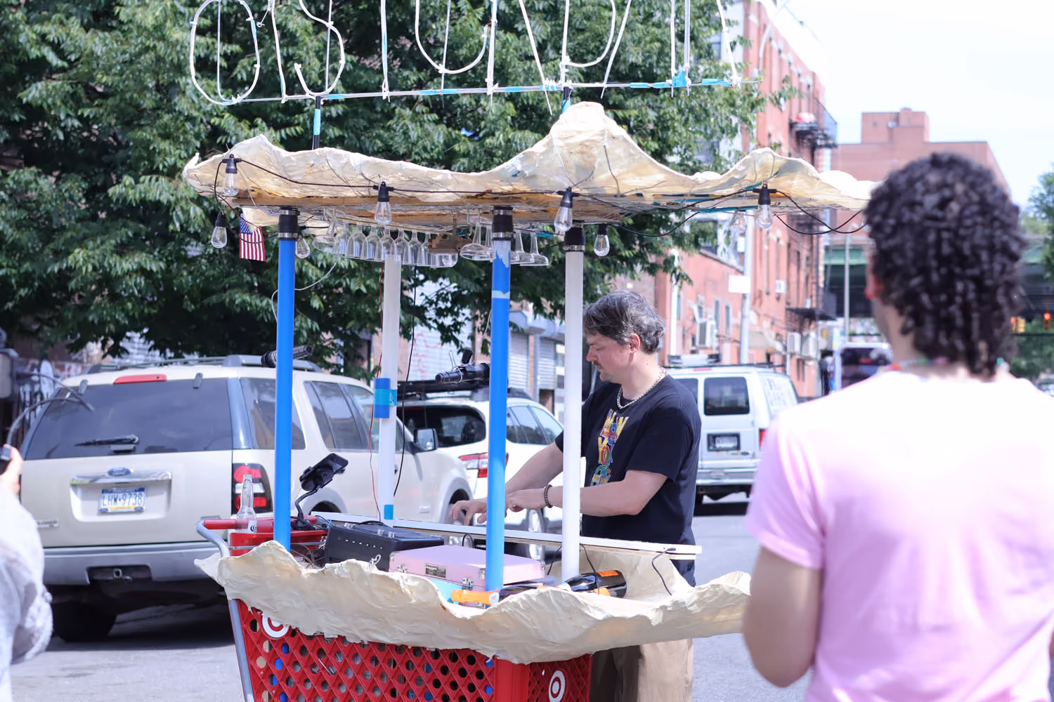 NARS Spring Open Studios 2026: a decorated cart with a canopy and blue poles on a Brooklyn street, with a person at the cart and a visitor in pink.