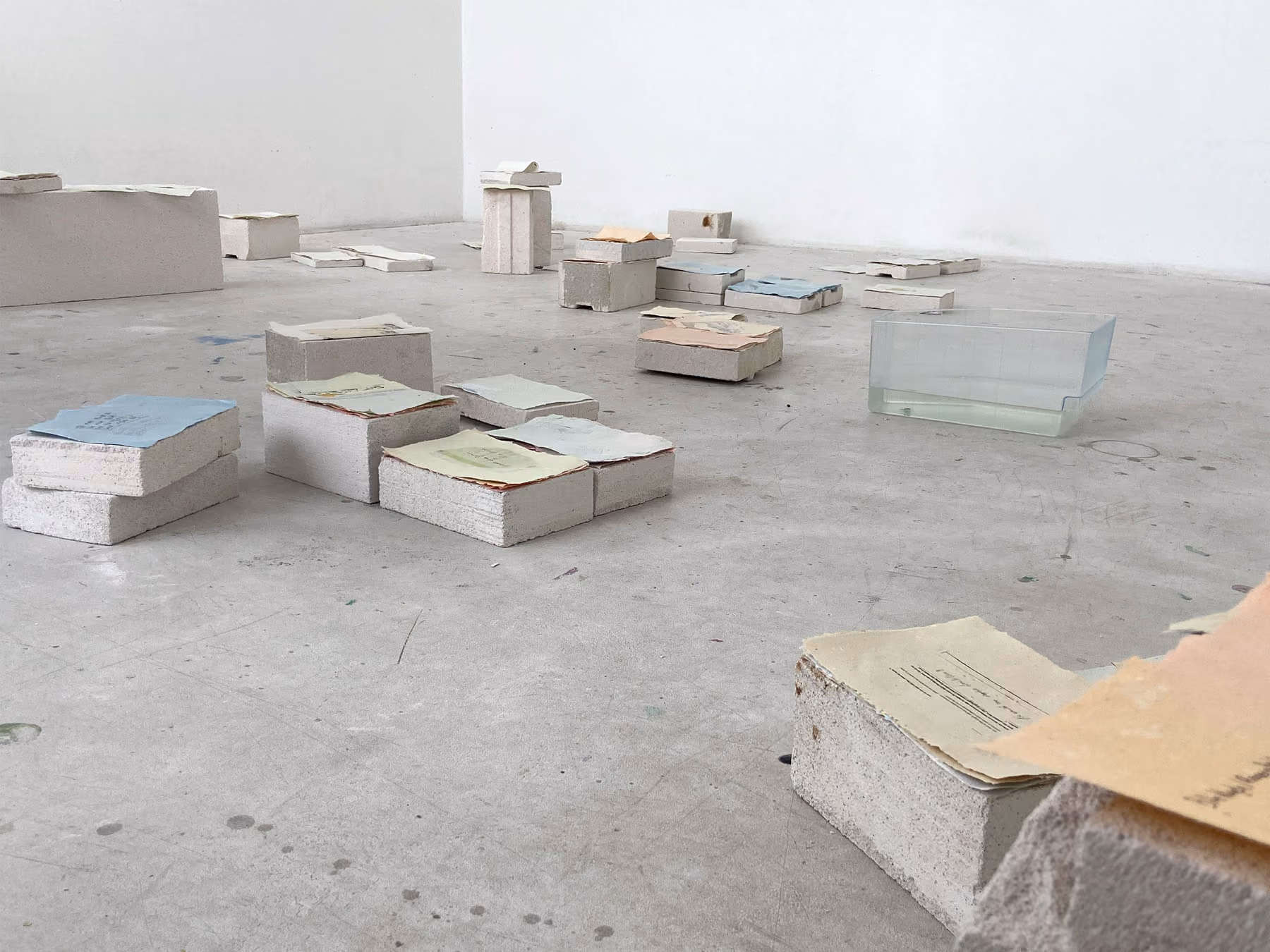 Pale concrete blocks topped with stacks of handmade paper in blue, green, and peach, scattered across a gray studio floor with a glass tank of water.