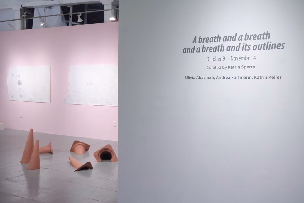 Photographed by Mashana Karimifar. Gallery entrance showing exhibition wall text reading "A breath and a breath and a breath and its outlines," curated by Katrin Sperry. In the background, terracotta-colored conical and funnel-shaped sculptures sit on the floor in front of a pink wall with hung paper works.