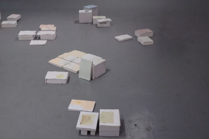 Photographed by Mashana Karimifar. High-angle view of various small, flat, rectangular blocks and stacks of pale pink, cream, and soft yellow paper sheets scattered across a smooth grey gallery floor.