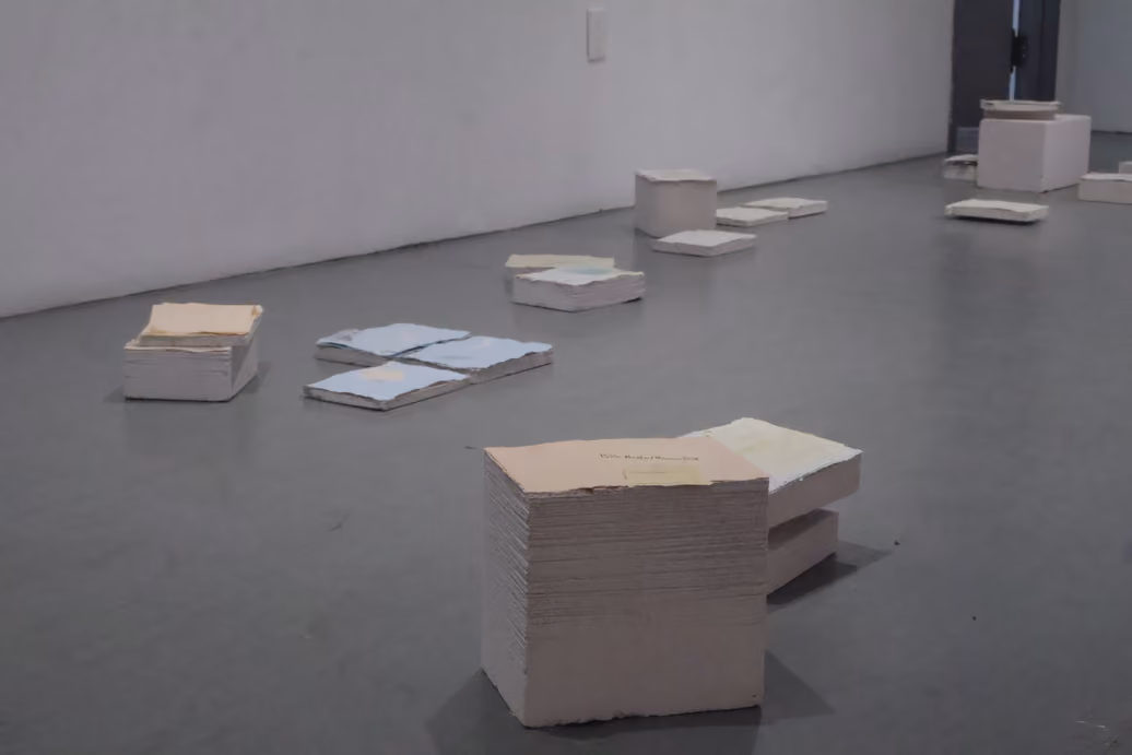 IMG_8586_26.jpg: Photographed by Mashana Karimifar. Art installation view featuring multiple stacks and scattered piles of thick, pastel-colored papers and block-like materials arranged across a polished grey gallery floor. White walls recede into the background.
