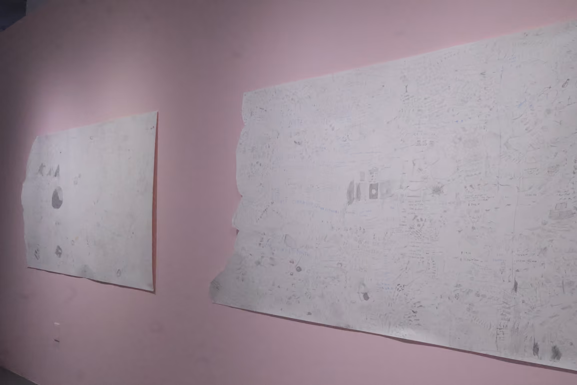 Photographed by Mashana Karimifar. Two large, light-colored paper sheets hanging on a light pink gallery wall. The sheets are covered with intricate, faint pencil drawings, diagrams, and handwritten notes.