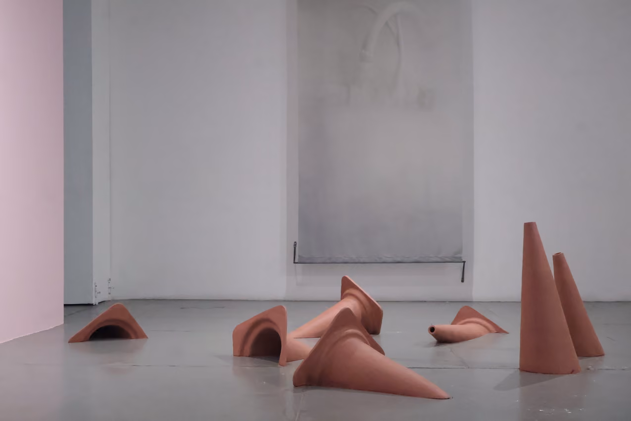 Photographed by Mashana Karimifar. Terracotta-colored ceramic cones and funnel-like sculptural pieces scattered across a grey floor in a gallery. In the background, a large vertical grey canvas hangs against a white wall alongside a pink corner wall.