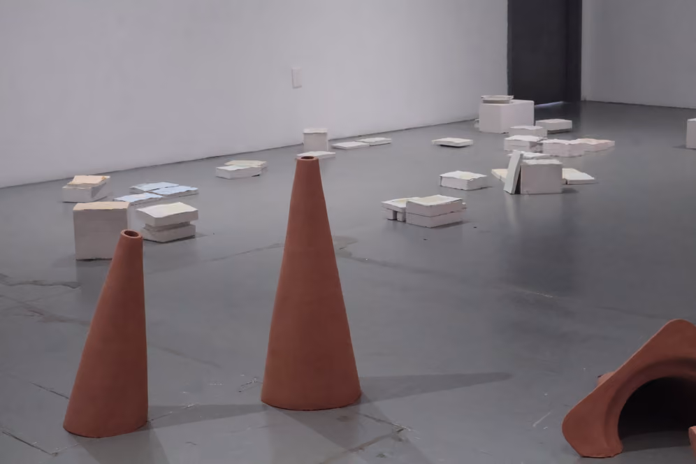 Photographed by Mashana Karimifar. Low-angle gallery view showing two upright terracotta cone sculptures in the foreground on a smooth grey floor. In the background, numerous stacks and flat arrangements of light pastel paper blocks extend toward a plain white wall.