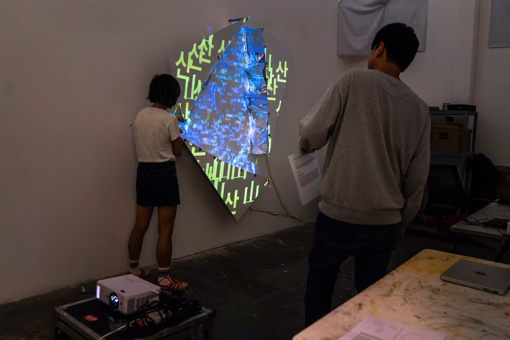 Photographed by Wei Chao. Two visitors interacting with a multimedia installation featuring green text and geometric light projections cast onto an angled wall surface.