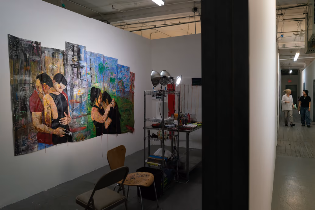 Photographed by Wei Chao. View into an open artist studio displaying a large multi-panel painted canvas of hugging figures alongside worktables and studio lighting. People walk down the hallway in the distance.
