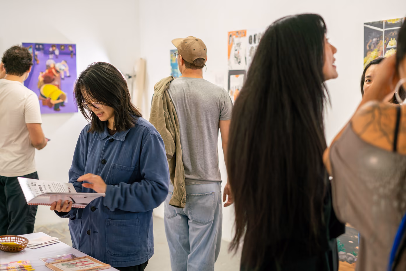 Photographed by Wei Chao. A visitor in a blue jacket flipping through an art publication at a display table surrounded by other gallery attendees.
