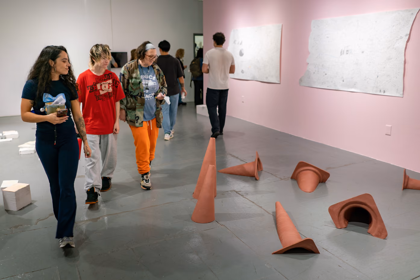 Photographed by Wei Chao. A group of visitors walking past terracotta cone sculptures scattered on the gallery floor next to large paper works on a pink wall.