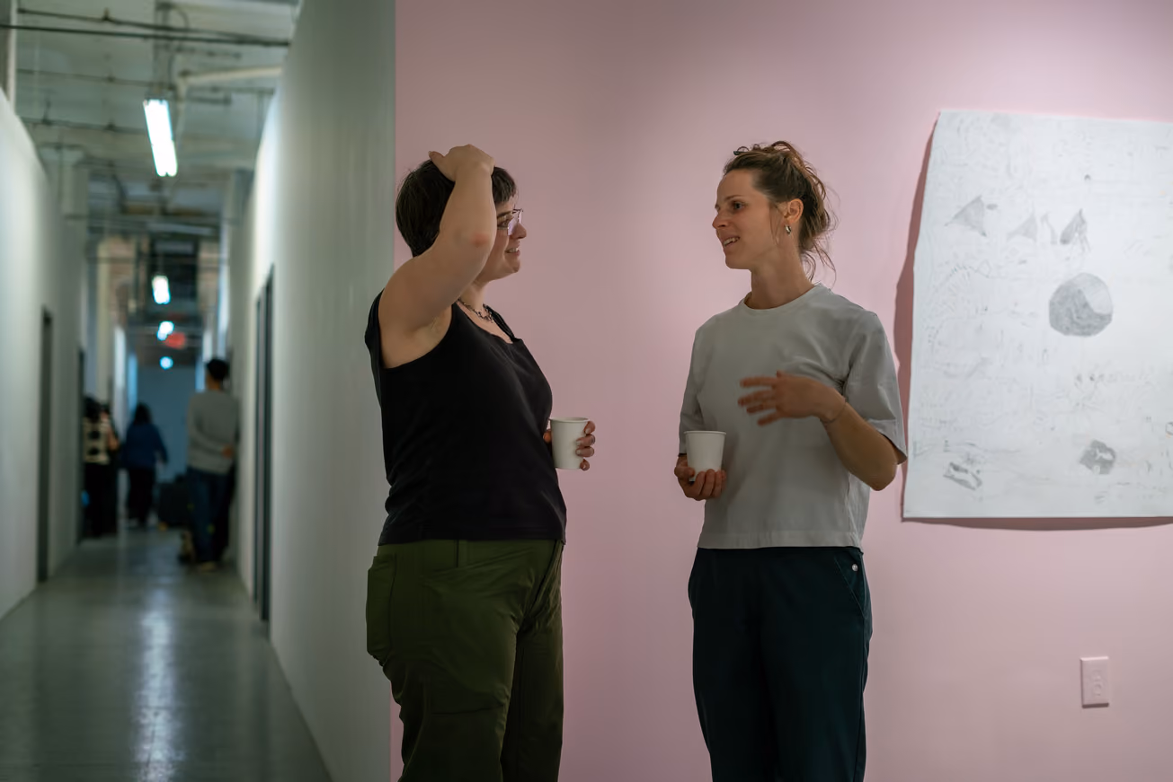 Photographed by Wei Chao. Two women holding paper cups and chatting in front of a pink wall with a large pencil drawing, situated beside a gallery hallway.